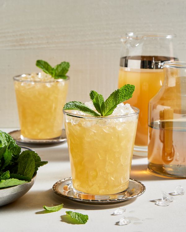 Deux verres de julep à la menthe sans alcool avec de la glace pilée et garnis de menthe, présentés avec un bol de feuilles de menthe, une carafe de julep préparé et une carafe de sirop simple à la menthe.