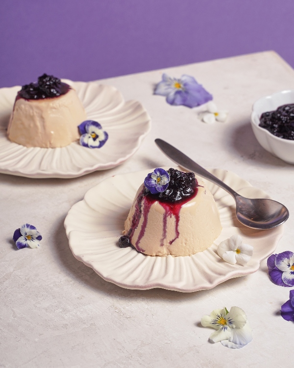 Deux portions de panna cotta à l'Earl Grey garnies de compote de bleuets et de fleurs comestibles, servies sur des assiettes crème à bord festonné avec une cuillère, sur un fond violet et crème doux.