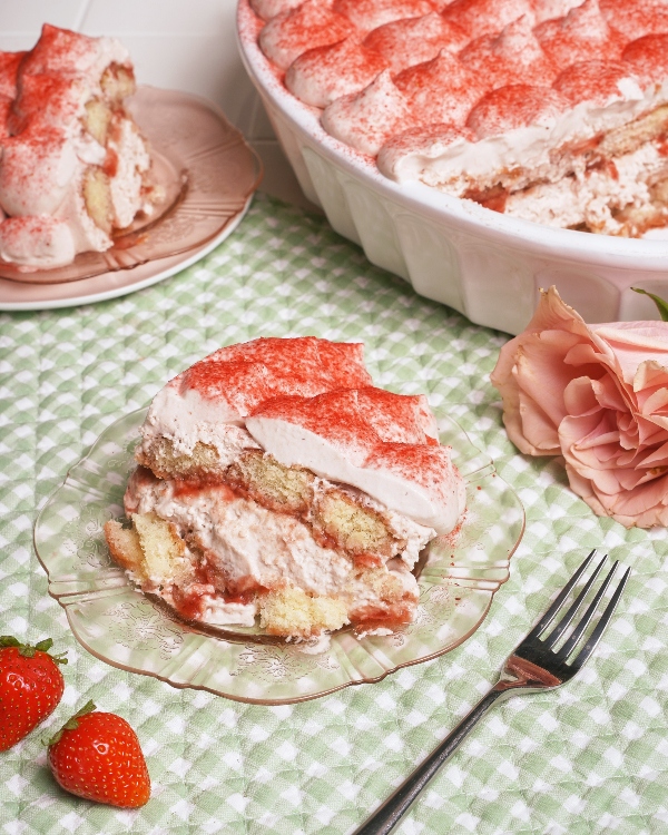 Un plat blanc contenant un tiramisu aux fraises et à la crème repose sur un tissu vichy vert, sur une surface en carreaux blancs. Deux portions coupées sont présentées sur des assiettes roses et en verre clair, avec des fraises fraîches et des roses roses tout autour.