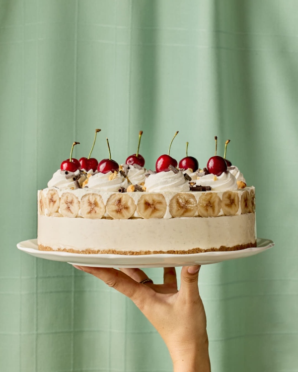 Une main tient une tarte glacée façon banana split sur une assiette blanche devant un arrière-plan vert menthe pâle. La tarte présente une croûte aux biscuits Graham, une garniture glacée crémeuse, un anneau de rondelles de banane sur les côtés et est couronnée de tourbillons de crème fouettée, de cerises au marasquin, de noix concassées et de copeaux de chocolat.