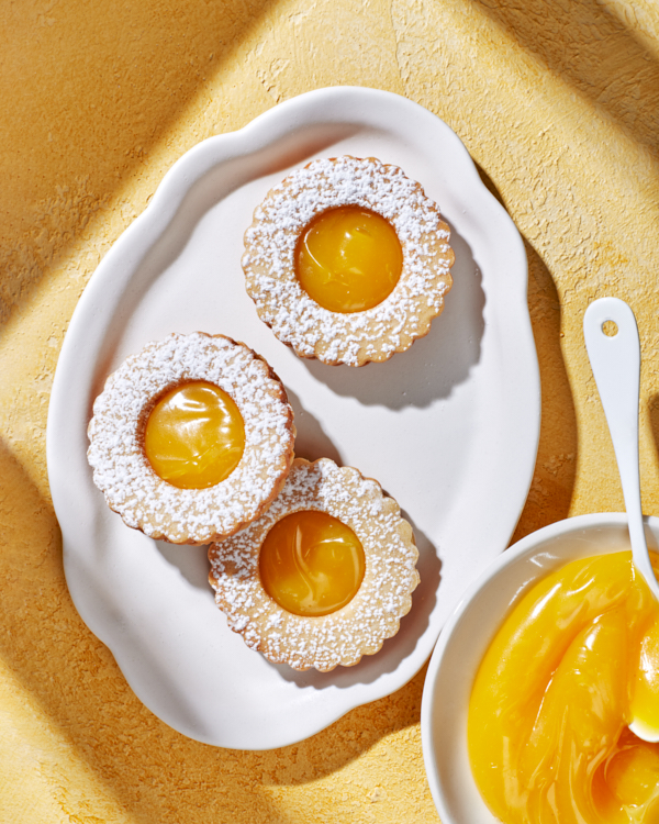 Biscuits sandwichs à la crème d'agrumes garnis de sucre glace sur un plateau blanc sur une surface jaune, montrés avec un bol de crème d'agrumes