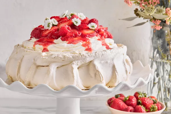 Une Pavlova garnie de framboises, de fraises et de fleurs sur un support à gâteau blanc, présentée avec un vase de fleurs et un bol de baies.