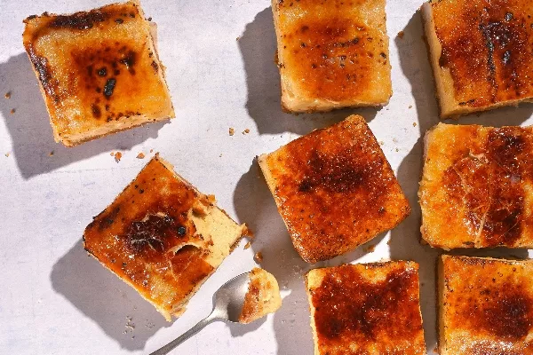 Une prise de vue de dessus de plusieurs petites tranches carrées de gâteau au fromage caramélisé avec une garniture de sucre brûlé, éparpillées sur une surface blanche avec une cuillère prenant une bouchée d'un morceau.