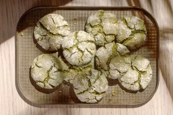 Une fournée de biscuits craquelés au matcha fraîchement sortis du four qui refroidissent sur une plaque à biscuits, montrant leurs surfaces craquelées et leur enrobage de sucre en poudre.