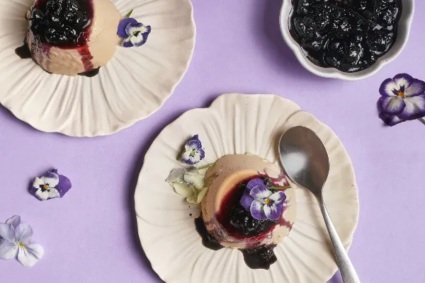Vue de dessus de deux portions de panna cotta à l'Earl Grey garnies de compote de bleuets et décorées de fleurs comestibles violettes et blanches, servies sur des assiettes crème à bord festonné sur un fond lavande avec des fleurs éparpillées et une cuillère.