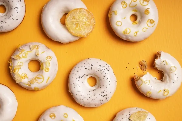 Des beignes glacés au citron et aux graines de pavot disposés sur un fond jaune vif, décorés de zeste de citron, de tranches de citron confit et de graines de pavot; une beigne a été croquée.