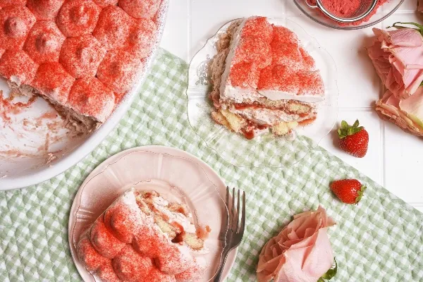 Un plat blanc contenant un tiramisu aux fraises et à la crème repose sur un tissu vichy vert, sur une surface en carreaux blancs. Deux portions coupées sont présentées sur des assiettes roses et en verre clair, avec des fraises fraîches et des roses roses tout autour.