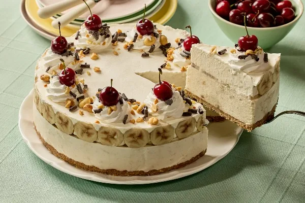 Une tarte glacée façon banana split couronnée de tourbillons de crème fouettée, de cerises, de noix concassées et de copeaux de chocolat déposée sur une assiette blanche. Une part est soulevée pour révéler des couches de rondelles de bananes et de garniture glacée sur une croûte de biscuits Graham. En arrière-plan, on voit une pile d’assiettes pastel avec des fourchettes et un bol de cerises fraîches sur une nappe vert pâle.