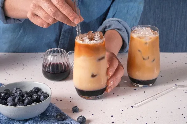  Une personne en chemise de jeans brasse un café glacé aux bleuets avec une paille en verre. La boisson a des couches distinctes de sirop de bleuets, de lait et de café. Une deuxième boisson identique est posée à proximité sur un comptoir moucheté. Un pichet en verre de sirop de bleuets, un bol de bleuets frais et des bleuets éparpillés complètent la scène.