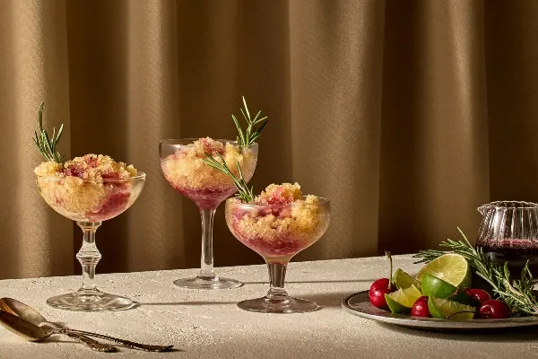 Trois coupes à cocktail remplies d’un dessert glacé composé de couches de slush dorée et rouge rubis, garnies de brins de romarin frais, posées sur une surface claire et texturée devant un rideau brun chaud. À côté, une assiette contient des limes entières, des quartiers de lime, des cerises et du romarin, ainsi qu’un petit pichet en verre rempli de sirop de cerise noire.