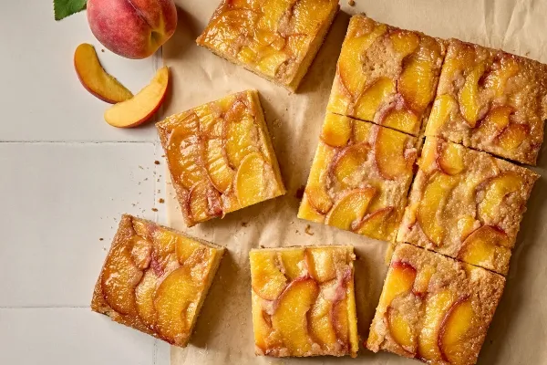 Vue plongeante de carrés renversés aux pêches bien dorés, placés sur un morceau de papier parchemin. Les carrés sont couronnés de tranches de pêches caramélisées lustrées. Une pêche fraîche entière et quelques quartiers de pêche sont disposés de manière décorative à côté des morceaux de dessert.
