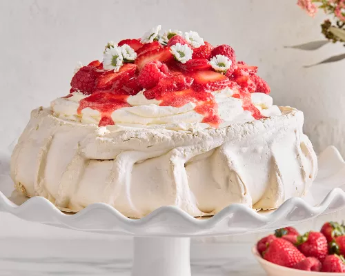 Une Pavlova garnie de framboises, de fraises et de fleurs sur un support à gâteau blanc, présentée avec un vase de fleurs et un bol de baies.