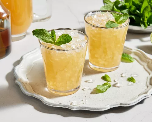 Deux verres de julep à la menthe sans alcool avec de la glace pilée et garnis de menthe, servis sur un plateau blanc et présentés avec un bol de feuilles de menthe, une moitié de lime, une carafe de julep préparé et une carafe de sirop simple à la menthe