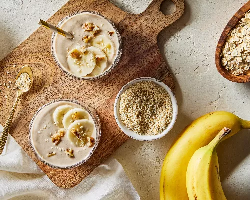  Vue de dessus de deux grands verres de smoothie à la banane, avoine et tahini, présentés sur une planche à découper en bois avec des bols de sucre jaune doré, de graines de sésame et d'avoine sèche, accompagnés de deux bananes non pelées.
