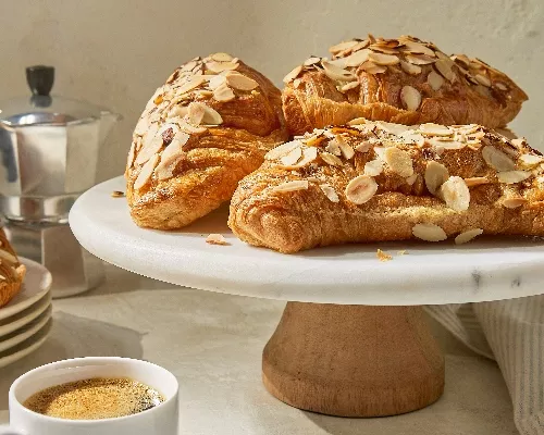 Quatre croissants aux amandes cuits à la friteuse à air, garnis d'amandes tranchées sur un présentoir à gâteaux blanc, avec une tasse d'espresso et une cafetière à l'arrière-plan.