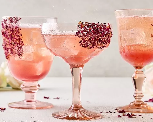 Trois verres à tige uniques de spritz litchi et rose sur glaçons. Des pétales de rose séchés garnissent les verres qui sont présentés dans un cadre neutre avec des roses fraîches en arrière-plan et des pétales de rose séchés dispersés autour.